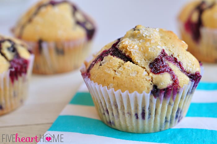 Classic Blueberry Muffins ~ these tender muffins are the BEST...bursting with berries and easy to make for a yummy breakfast or snack! | FiveHeartHome.com Close-up of Blueberry Muffins in paper wrappers, sitting on light blue and white striped napkinds