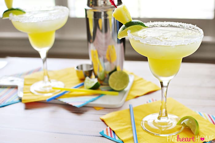 Two garnished glasses on table with shaker in background.