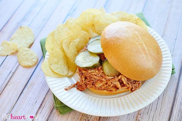 Chicken Sloppy Joe in a bun, topped with sliced pickles and on a plate with potato chips.