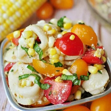 Tortellini Pasta Salad in a bowl with corn on the cob, colorful grape tomatoes, and basil leaf in background