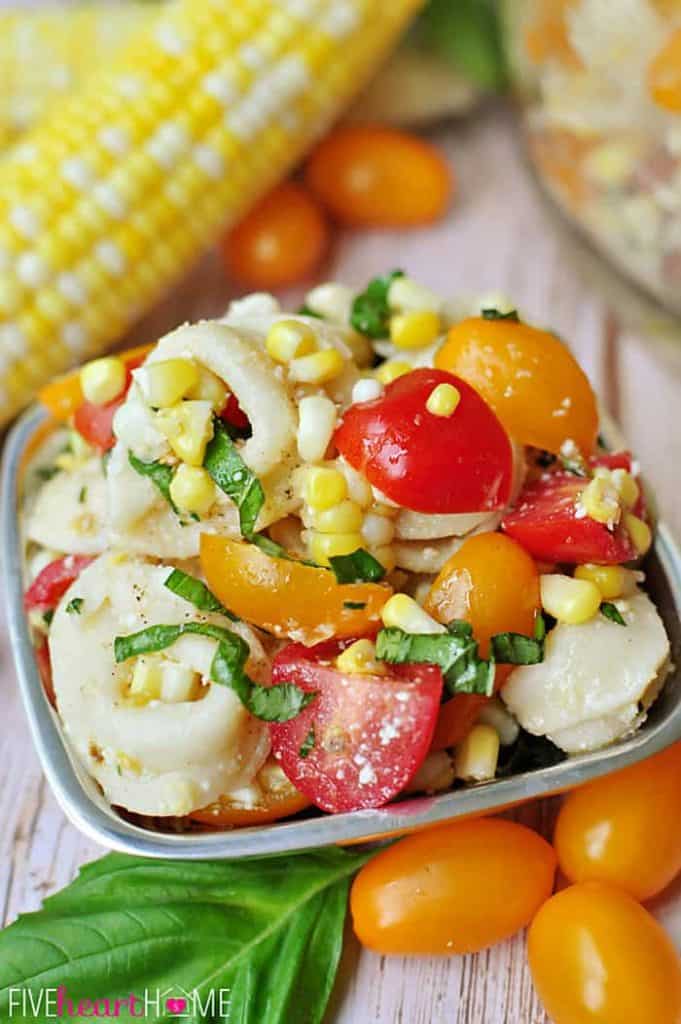 Tortellini Pasta Salad in a bowl with corn on the cob, colorful grape tomatoes, and basil leaf in background