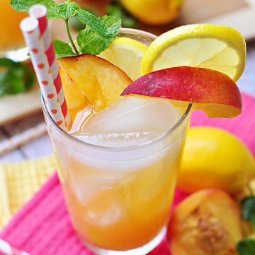 Glass of peach lemonade garnished with fresh peach wedge, lemon slice, sprig of mint, and two red and white paper straws