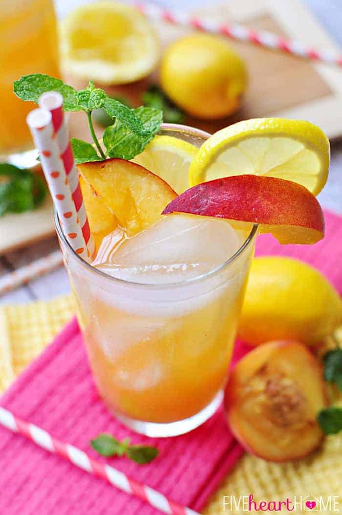 Glass of peach lemonade garnished with fresh peach wedge, lemon slice, sprig of mint, and two red and white paper straws