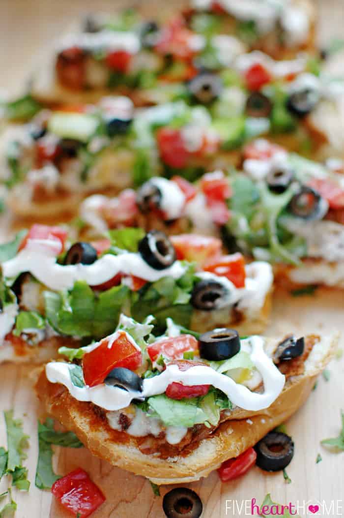 French Bread Taco Pizza ~ tacos meet pizza on an effortless French bread crust, with layers of refried beans, salsa, taco meat, cheese, lettuce, tomatoes, black olives, and sour cream! | FiveHeartHome.com French Bread Taco Pizza Sliced on a Cutting Board with All the Fixings