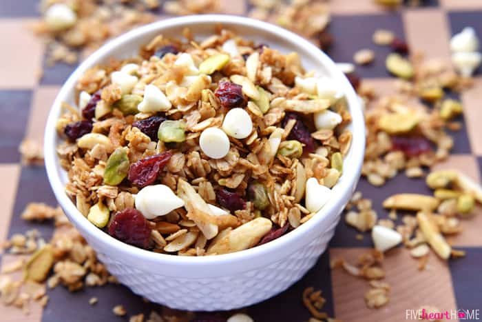 Gingerbread Granola with Cranberries, Pistachios, and White Chocolate Chips ~ warm spices and Christmas colors make this a perfect holiday breakfast or homemade food gift! | FiveHeartHome.com Overflowing bowl with extra scattered on table.