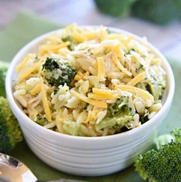 Broccoli Cheese Orzo with Chicken in a white bowl with a spoon and broccoli scattered on table