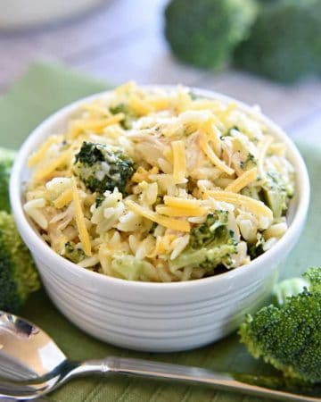 Broccoli Cheese Orzo with Chicken in a white bowl with a spoon and broccoli scattered on table