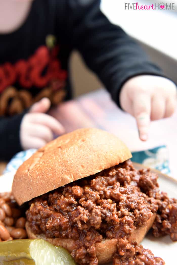 Toddler trying to touch a sloppy joes sandwich on a plate.