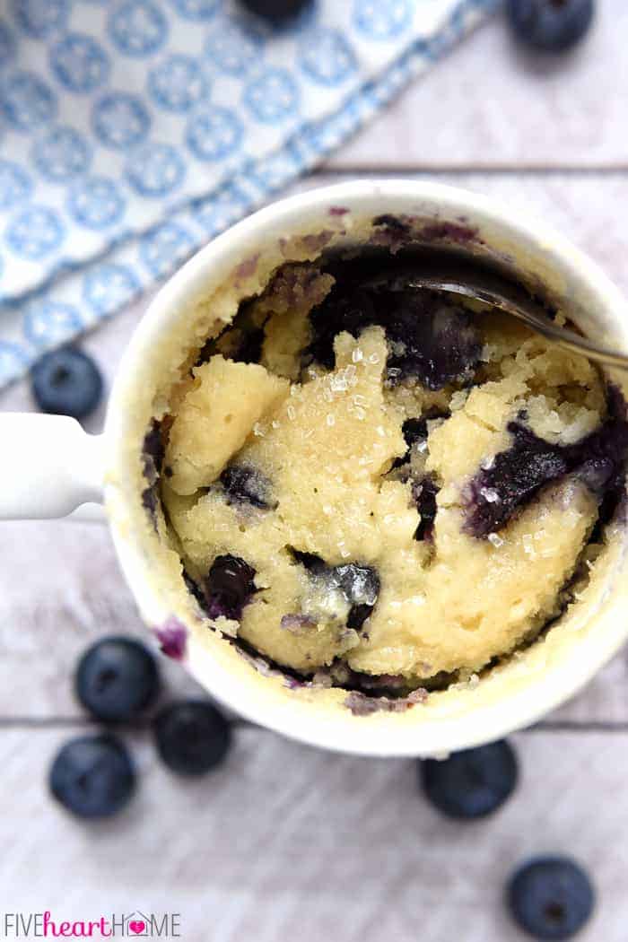 Aerial view of Blueberry Muffin Mug Cake with fresh blueberries and blue and white towel on table.