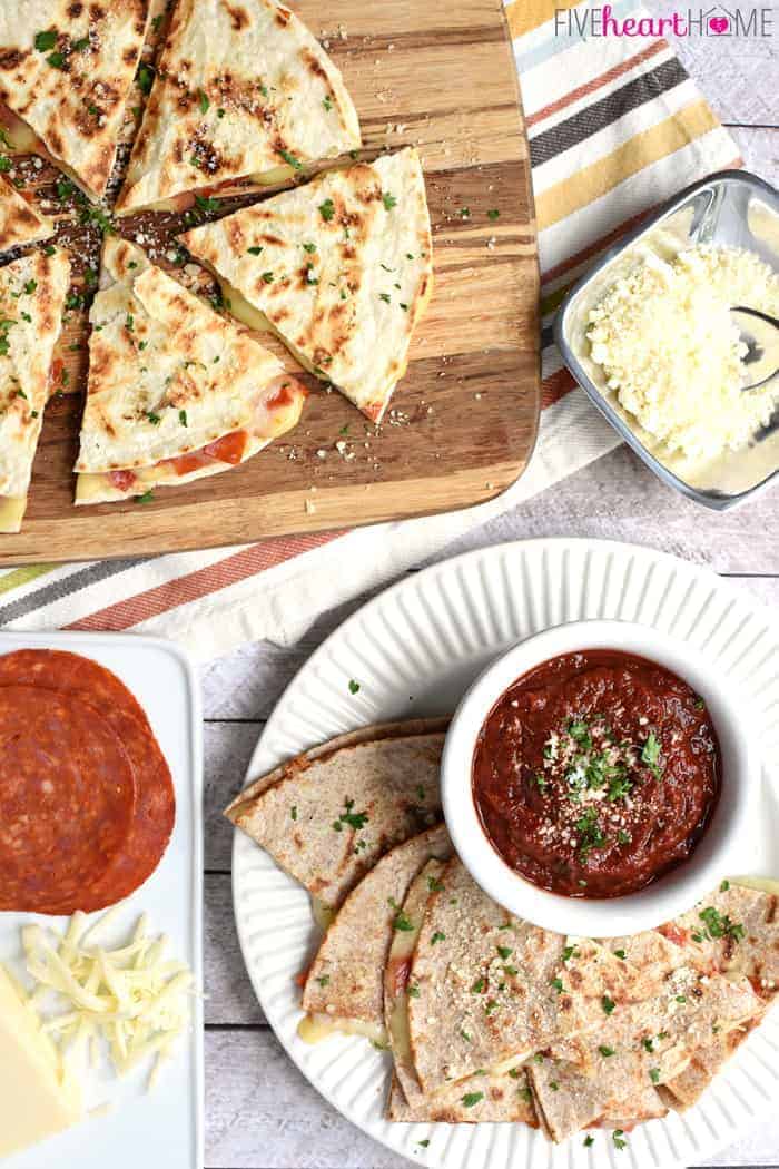 Aerial view of Pizza Quesadillas with Dipping Sauce on a plate, plus more sliced quesadillas on a cutting board, plus ingredients for making them scattered on the table
