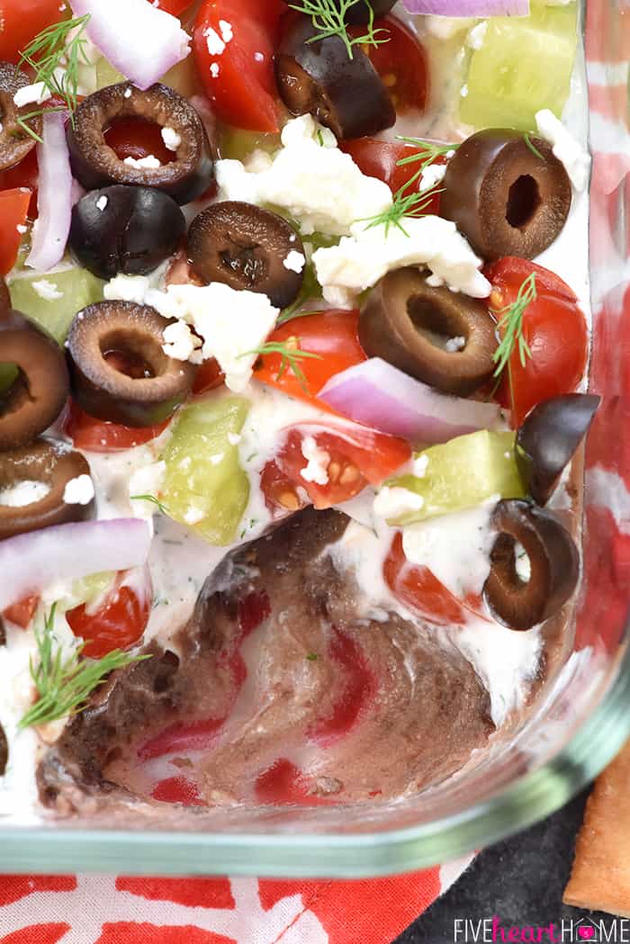 Greek 7-Layer Dip ~ a Mediterranean spin on the beloved, classic recipe, with layers of hummus, dill-infused Greek yogurt, tomatoes, cucumbers, black olives, red onion, and feta cheese! | FiveHeartHome.com Aerial view of dish of Greek 7-Layer Dip with missing scoop.