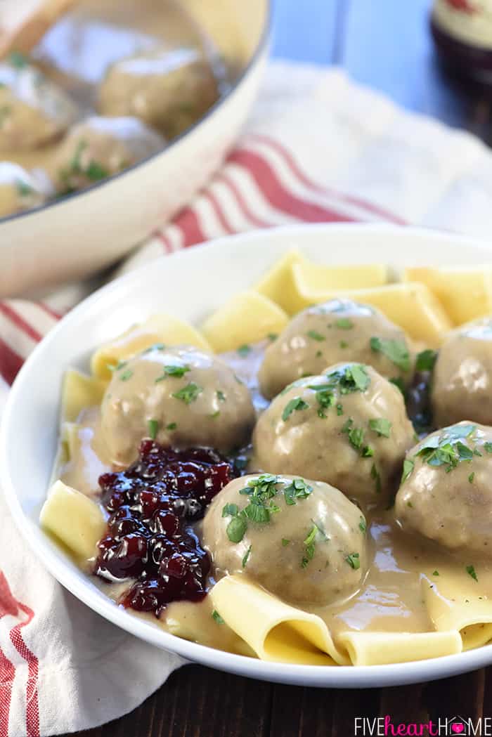 Plate of meatballs, gravy, lingonberry jelly, and parsley over egg noodles, with edge of skillet in background