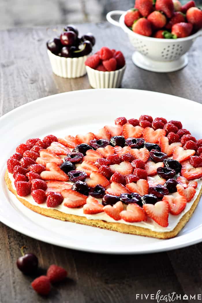 Fruit Pizza ~ on a heart-shaped sugar cookie crust, this fruit pizza features cream cheese frosting topped by glossy berries & cherries. It's a perfect Valentine's Day dessert or treat for any occasion! | FiveHeartHome.com.com Fruit Pizza on white platter with bowls of strawberries, raspberries, and cherries in background.