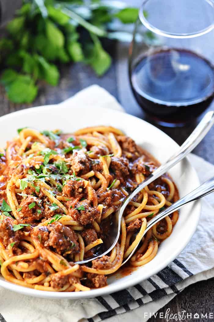 Pasta and meatsauce in a bowl with parsley and glass of wine in background.