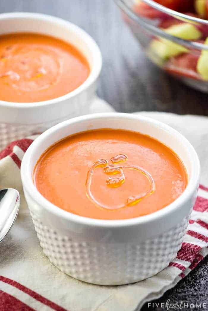 Two bowls of Easy Gazpacho recipe with bowl of chopped veggies in background.