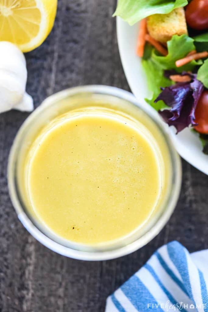 Aerial view of jar of Champagne Vinaigrette surrounded by head of garlic and salad on table.