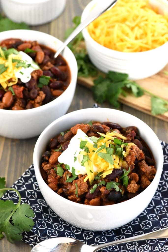 3 Bean Chili in two bowls with cheese, sour cream, and cilantro on top.