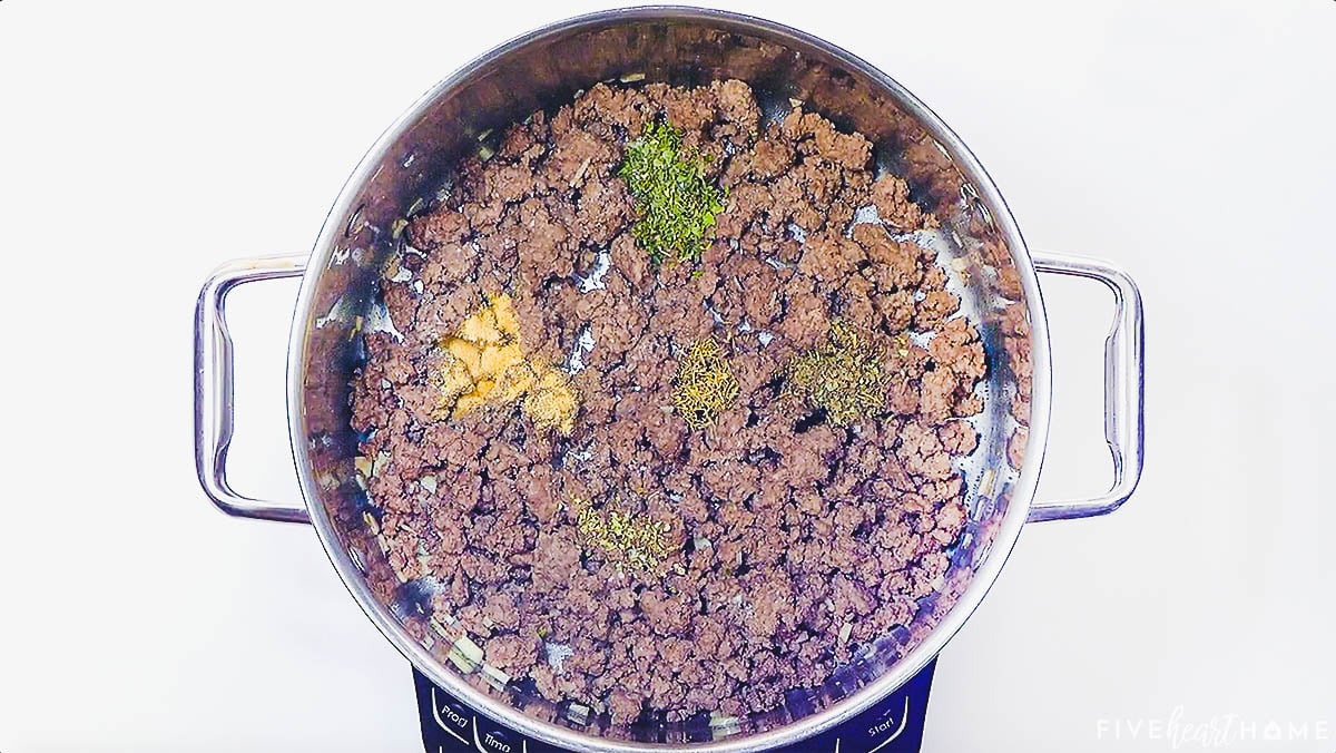 Seasoning ground beef in pot with herbs and spices.