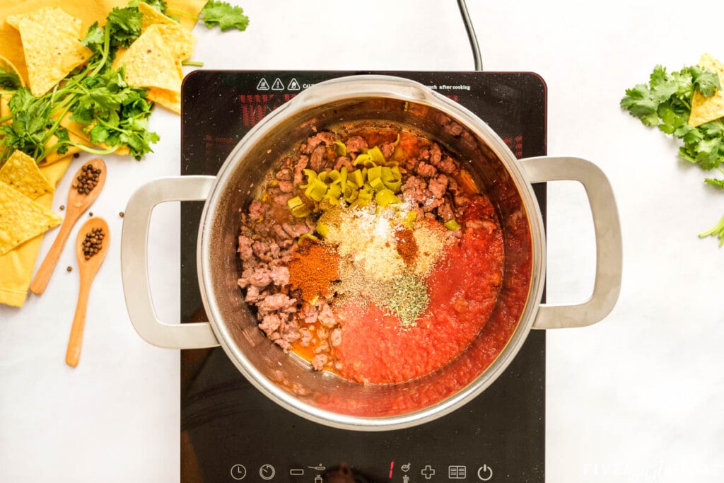 Adding tomato sauce, diced green chiles, and spices to cooked ground beef in pot for Enchilada Dip.