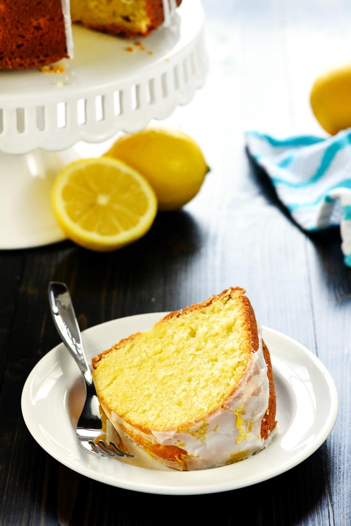 Slice of Lemon Pound Cake recipe on white plate with lemons in background.