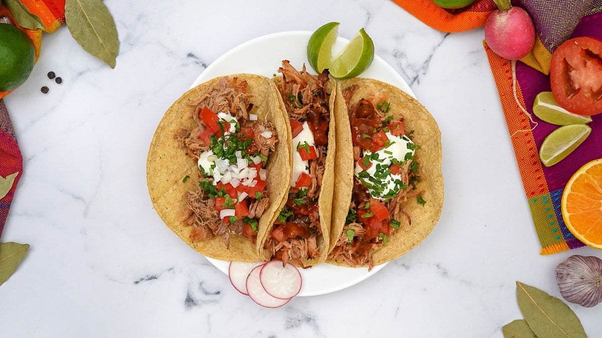 Aerial view of three Carnitas Tacos on plate with garnishes.