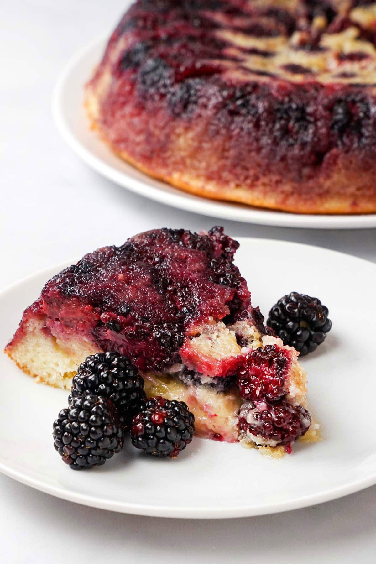 Blackberry Cake slice on plate with blackberry upside-down cake in background.