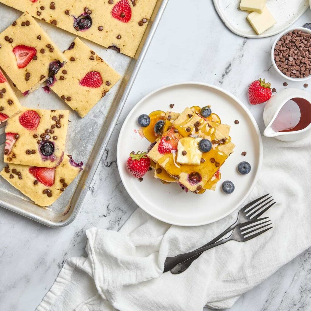 Sheet Pan Pancakes aerial view on plate with syrup and on sheet pan with berries and chocolate chips.